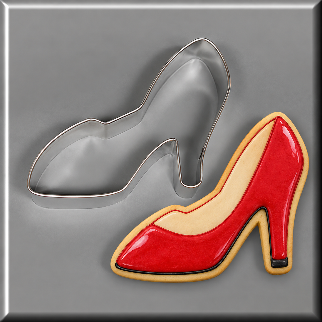 A red high-heeled shoe-shaped cookie with beige icing details and a black accent on the heel, shown next to a 4 inch Shoe or Slipper Metal Cookie Cutter on a gray background.