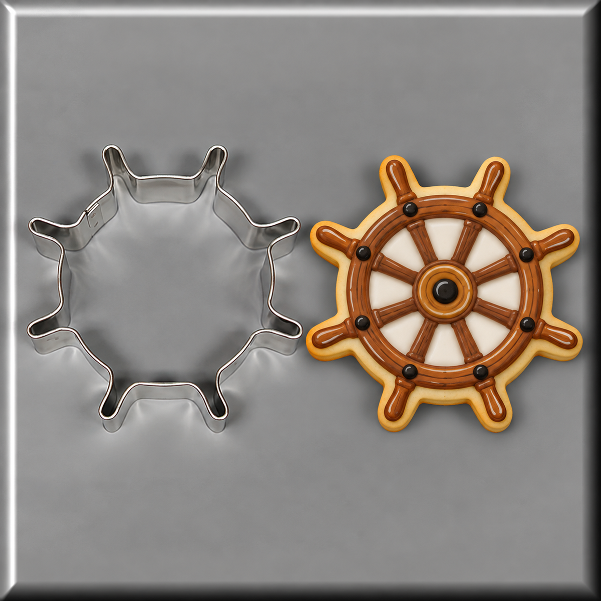 A ship's wheel shaped sugar cookie with brown icing and white sections, shown next to a 4 inch Ship's Wheel Metal Cookie Cutter on a gray background.