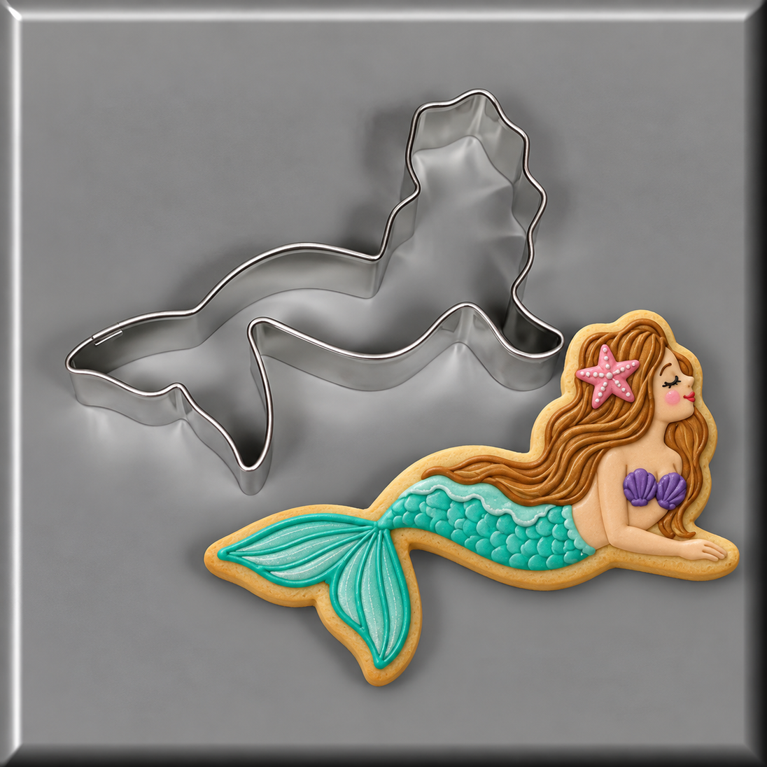 A 4 inch Mermaid Metal Cookie Cutter shown above a decorated sugar cookie of a mermaid with a turquoise tail, brown hair, and a purple shell, on a gray background.