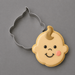 A baby-shaped sugar cookie with light beige icing, decorated with a smiling face, black eyes and mouth, a pink cheek, and a brown tuft of hair, shown next to a 4.25 inch Baby Face Metal Cookie Cutter on a gray background.