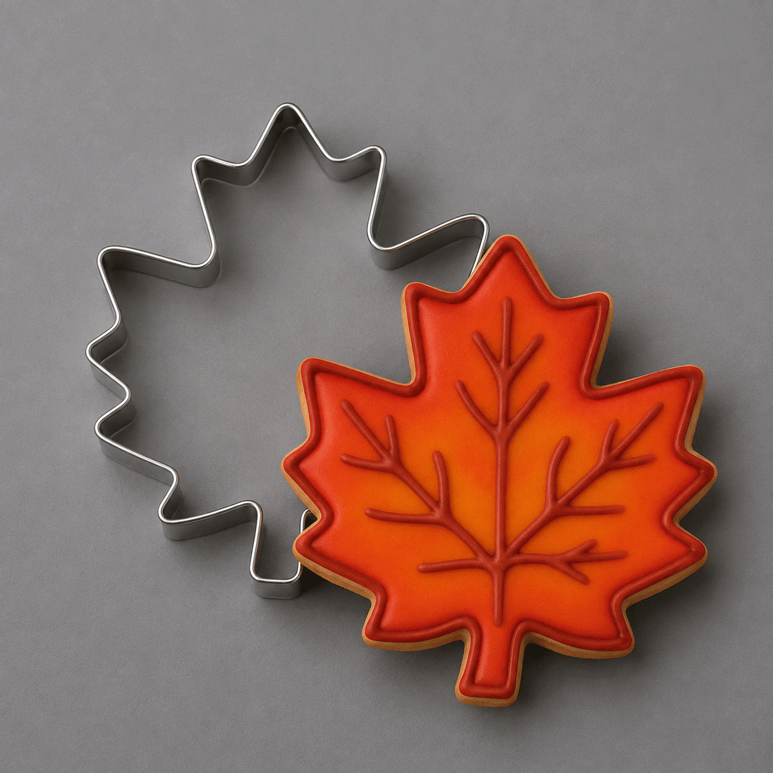 A metal maple leaf-shaped cookie cutter positioned behind a decorated maple leaf cookie with orange and red icing, set against a solid gray background from a 3.5 inch Maple Leaf Metal Cookie Cutter